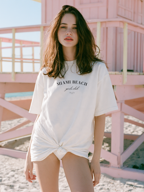 Woman wearing an ivory Miami Beach Girls Club oversized tee knotted at the hip in front of a pink lifeguard tower on Miami Beach