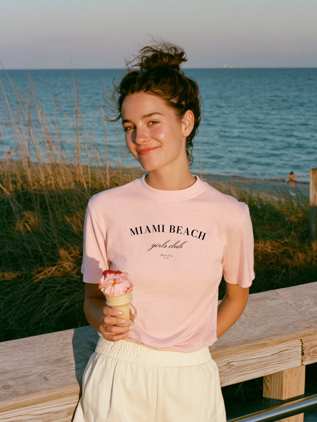 Woman holding an ice cream cone in a blossom pink Miami Beach Girls Club Comfort Colors tee with the ocean behind her