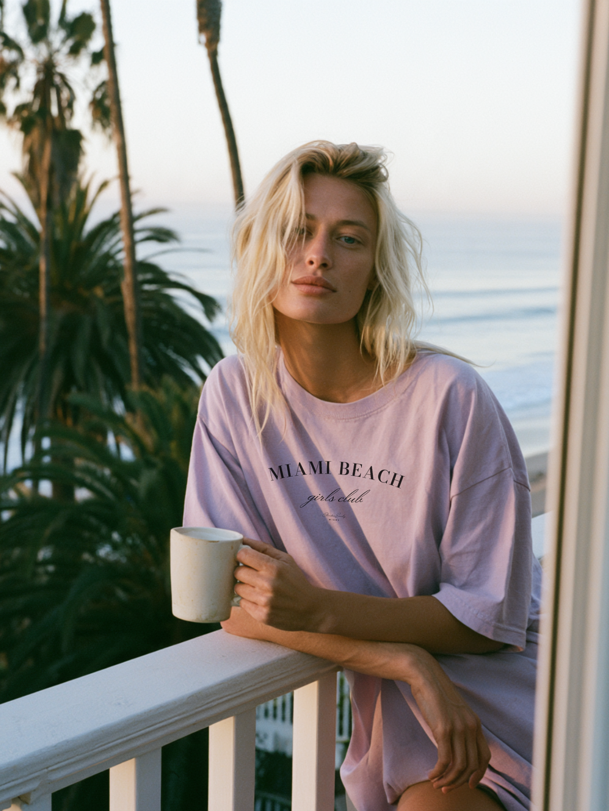 Blonde woman holding a coffee mug in an orchid purple Miami Beach Girls Club oversized tee on a balcony with palm trees and ocean views
