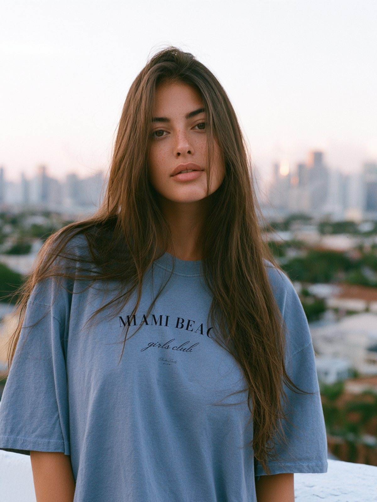 Woman in a chambray blue Miami Beach Girls Club oversized tee with the Miami skyline at sunset behind her